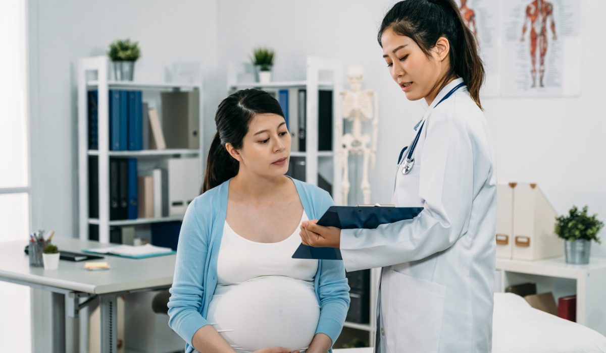 asian female physician standing with a clipboard is explaining diagnosis to her expectant patient who’s sitting on treatment bed in the hospital.